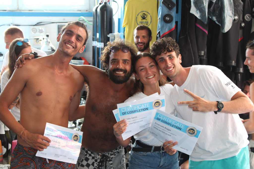 Giovani subacquei festeggiano il completamento del corso Open Water Diver con certificati in mano presso un diving center.

