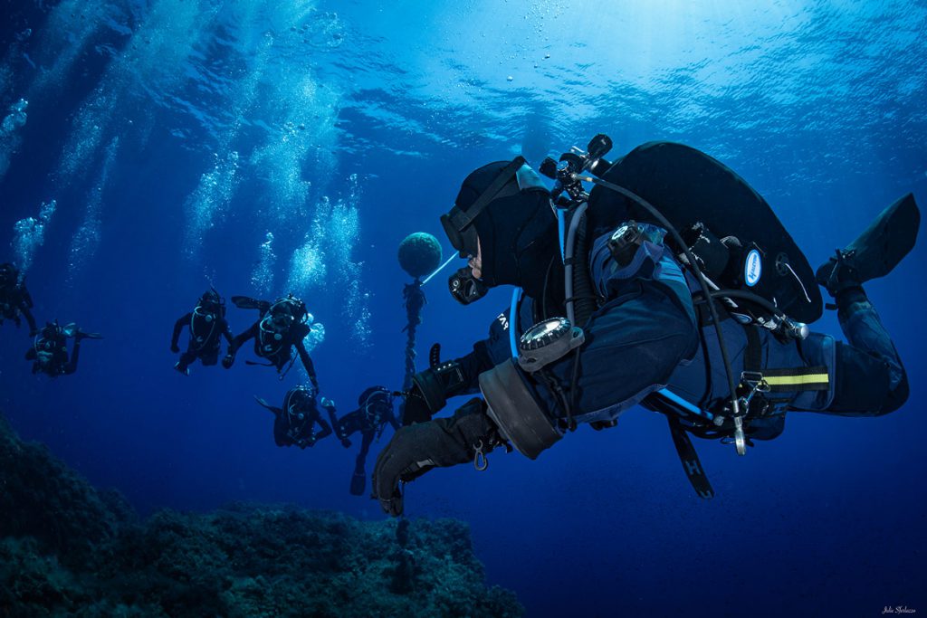 Gruppo di subacquei mentre effettua la discesa in acque profonde, equipaggiati con mute e bombole.
