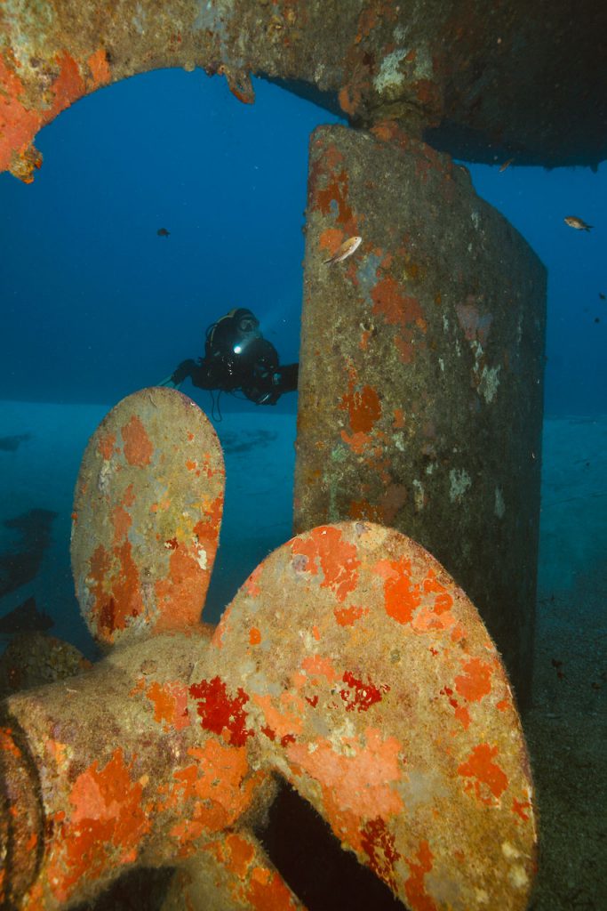 Vista ravvicinata dell'elica e del timone del relitto Um El Faroud a Malta, con subacqueo in immersione tecnica sullo sfondo