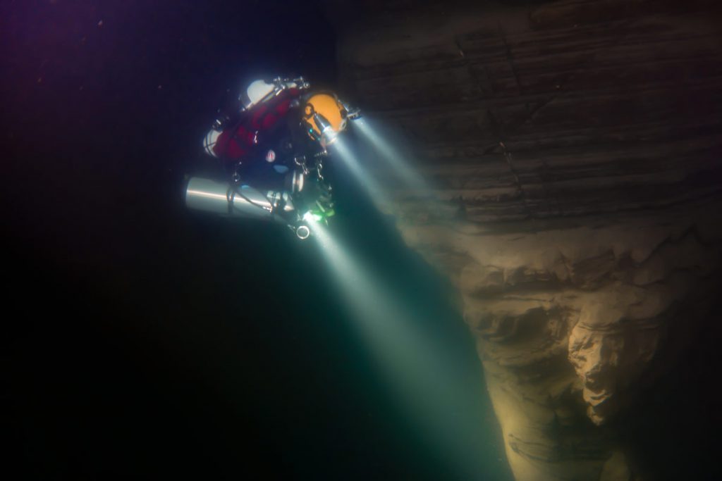 Subacqueo tecnico illumina una parete verticale stratificata nel Lago di Como durante un’immersione in profondità