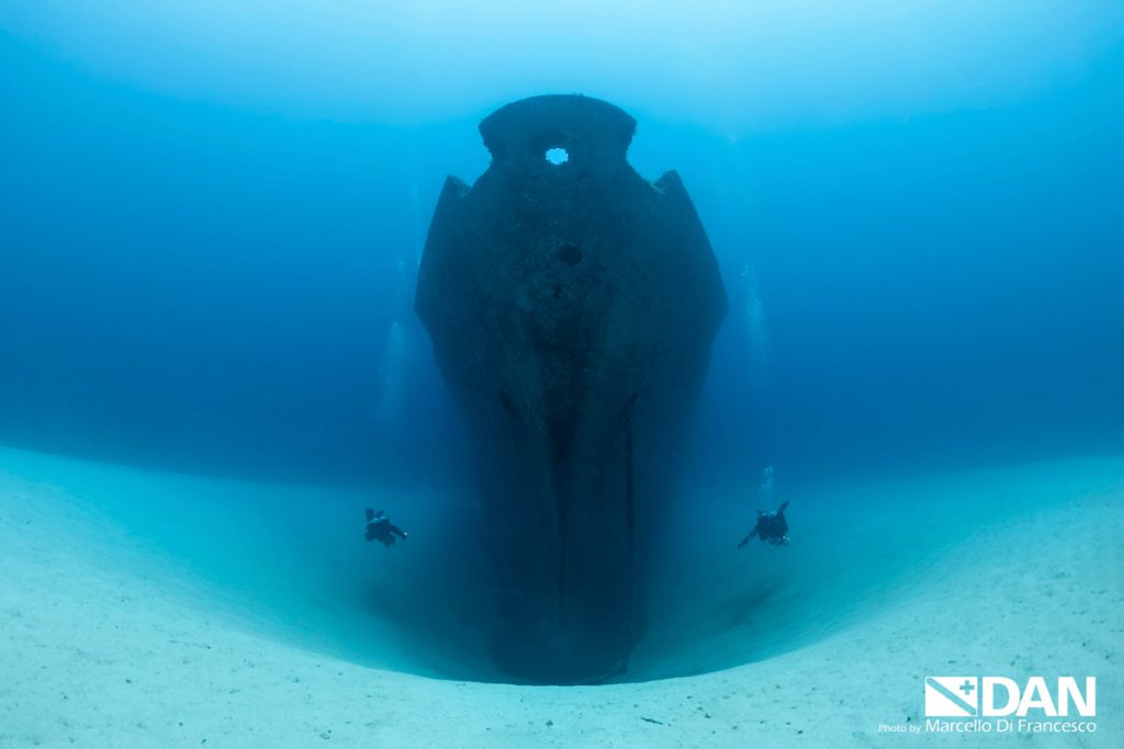 Imponente prua del relitto Um El Faroud a Malta con due subacquei in esplorazione sul fondale sabbioso