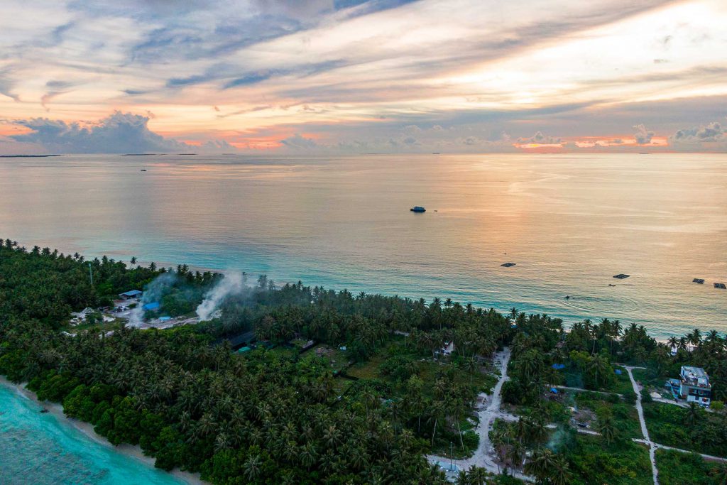 Vista aerea di un'isola delle Maldive con palme, edifici e fumo, affacciata su un mare calmo al tramonto